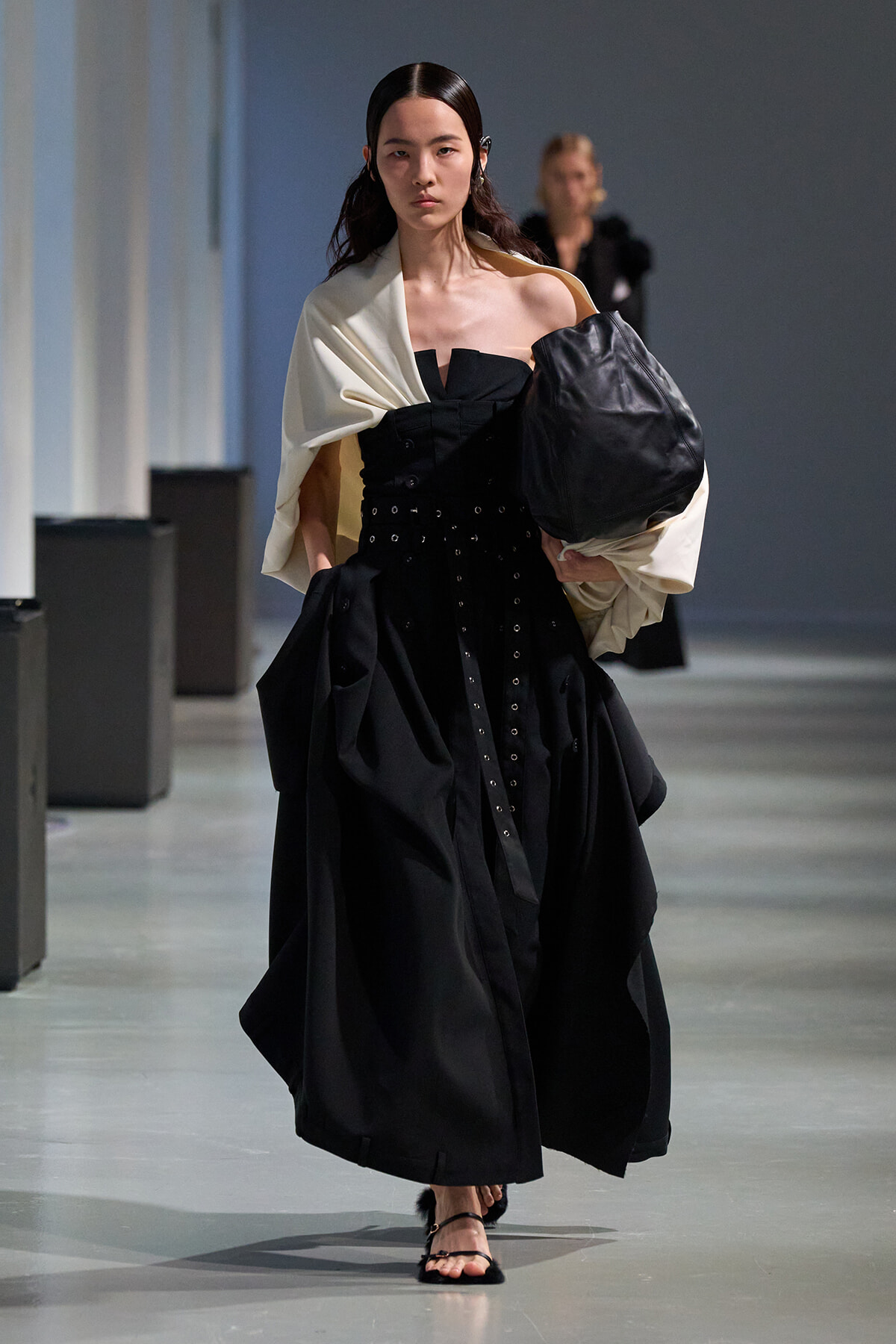 Model walking a fashion runway in a black strapless gown with a cream draped shawl and oversized black bag on her right arm, wearing sandals.