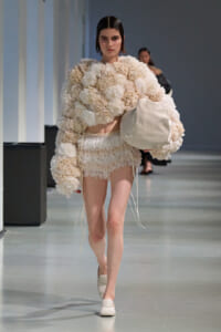 Model on a runway wearing a cream floral oversized top and matching ruffled shorts, carrying a large cream tote.