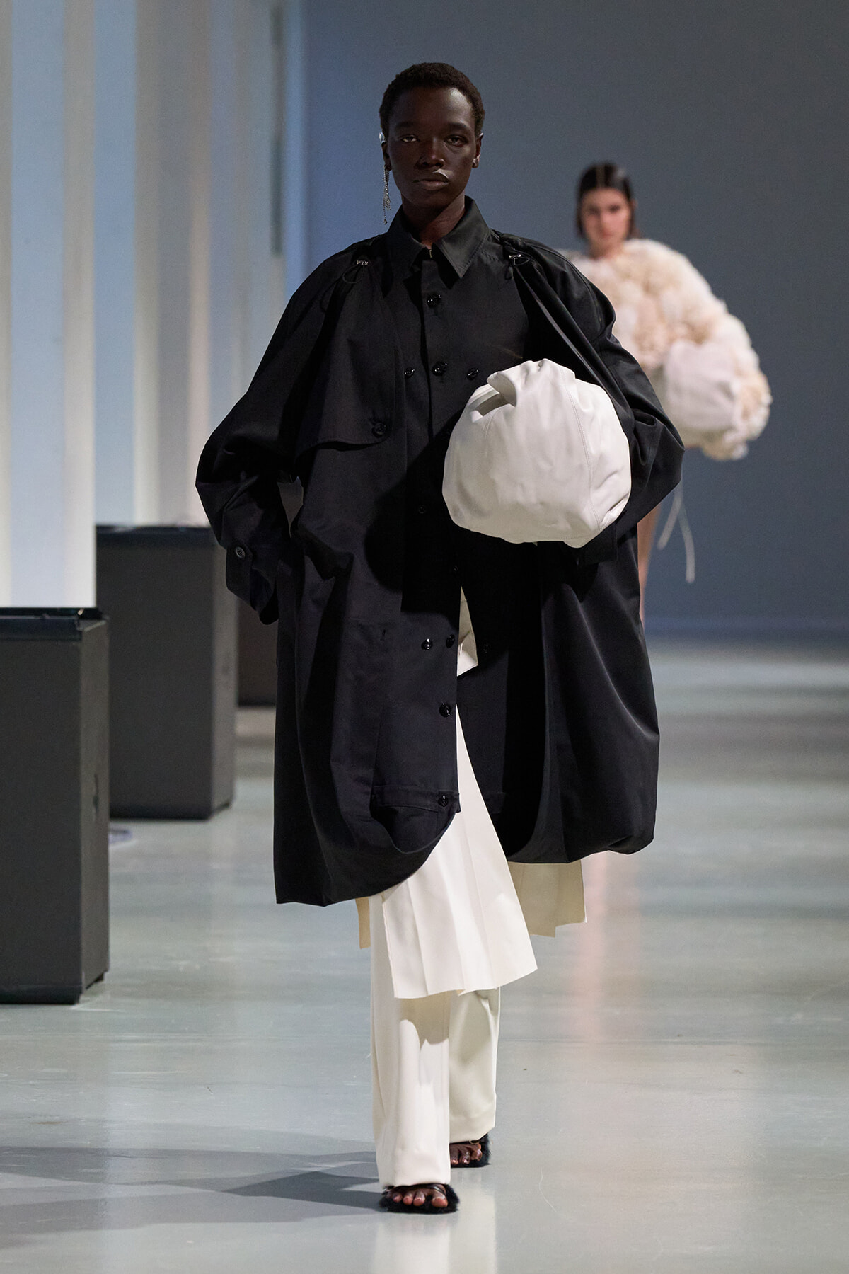 Model walking a fashion runway in an oversized black coat over white pants, carrying a white bag.