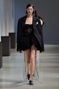 Model walking runway in an oversized black blazer over a sheer black ruffled mini dress with long fringe cords, black loafers, and metallic face jewelry.