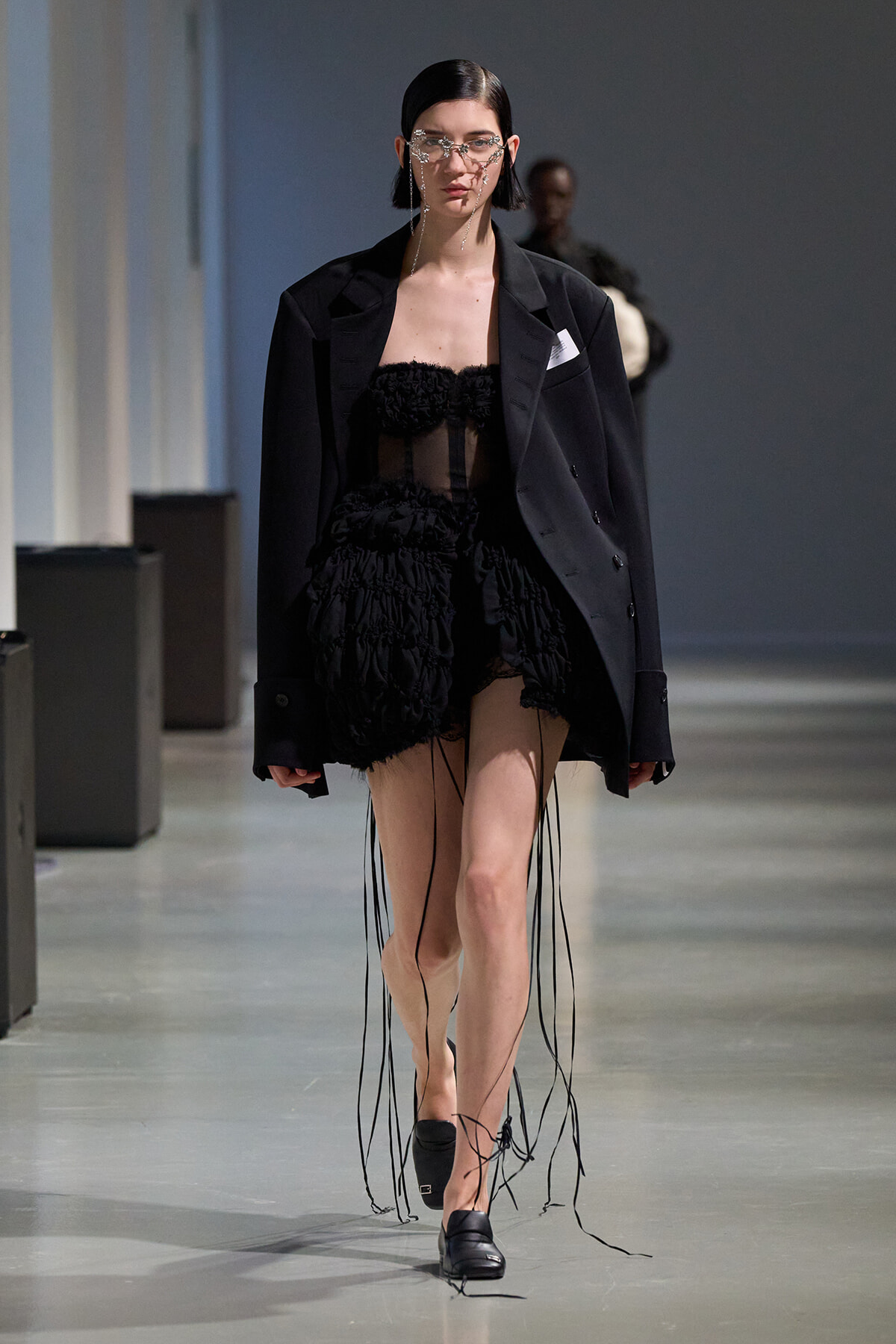 Model walking runway in an oversized black blazer over a sheer black ruffled mini dress with long fringe cords, black loafers, and metallic face jewelry.
