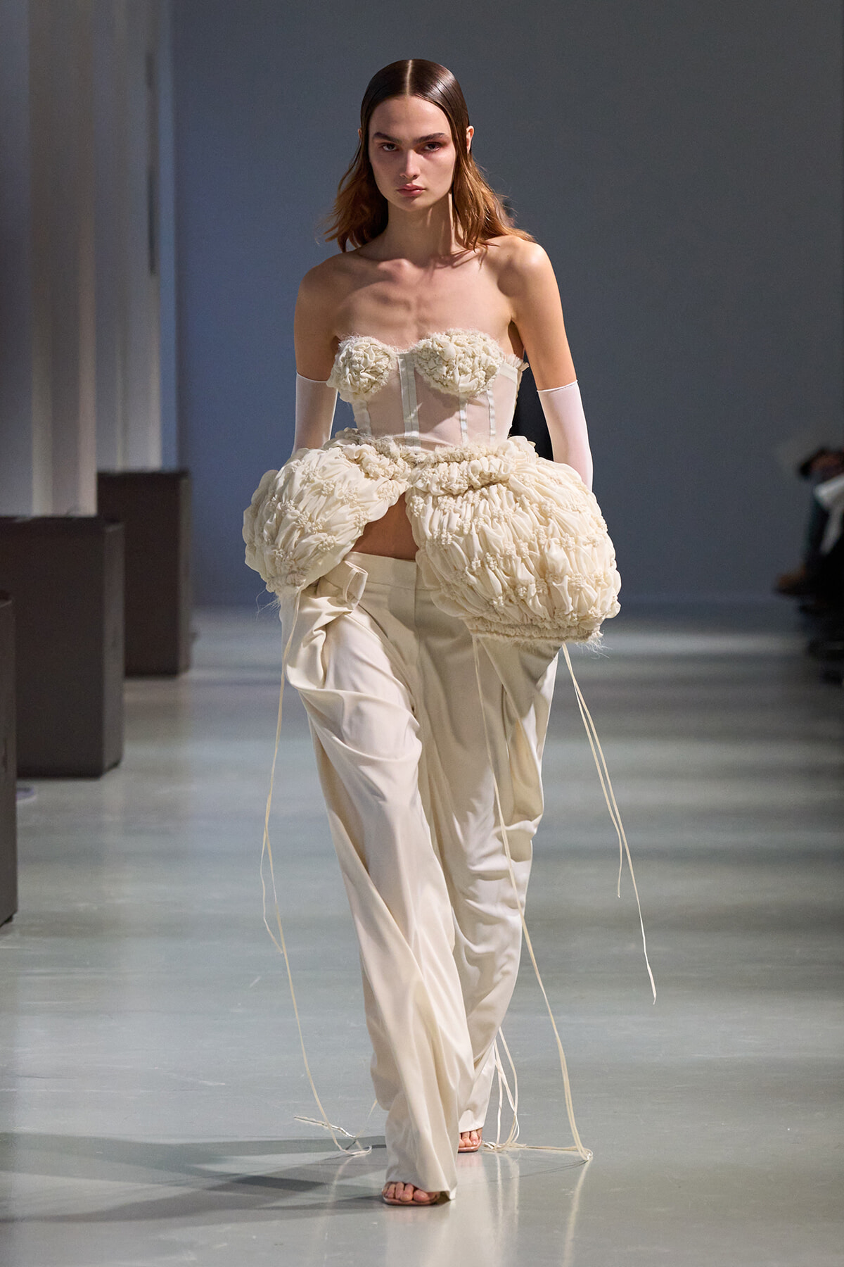 Model on a fashion runway wearing an ivory strapless corset with floral appliqués and a voluminous ruched skirt, pale gloves.
