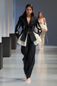 Model walks the runway in a black tailored blazer with white fabric panels and matching wide-leg pants.