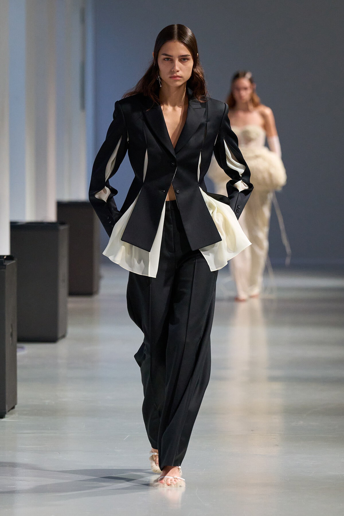 Model walks the runway in a black tailored blazer with white fabric panels and matching wide-leg pants.