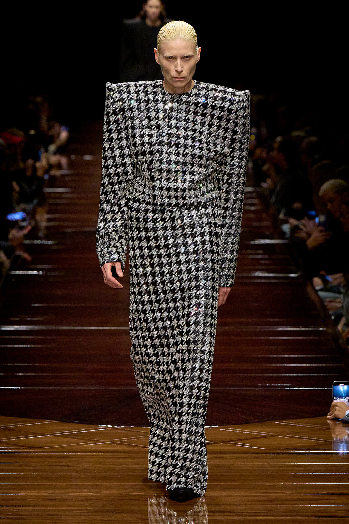 Model walking a fashion runway in a sequined black-and-white houndstooth suit with exaggerated shoulders and wide legs, slick blond hair.