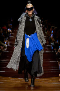 Model walks a fashion runway in a black outfit with a houndstooth coat, blue wrap skirt, and futuristic visor sunglasses.