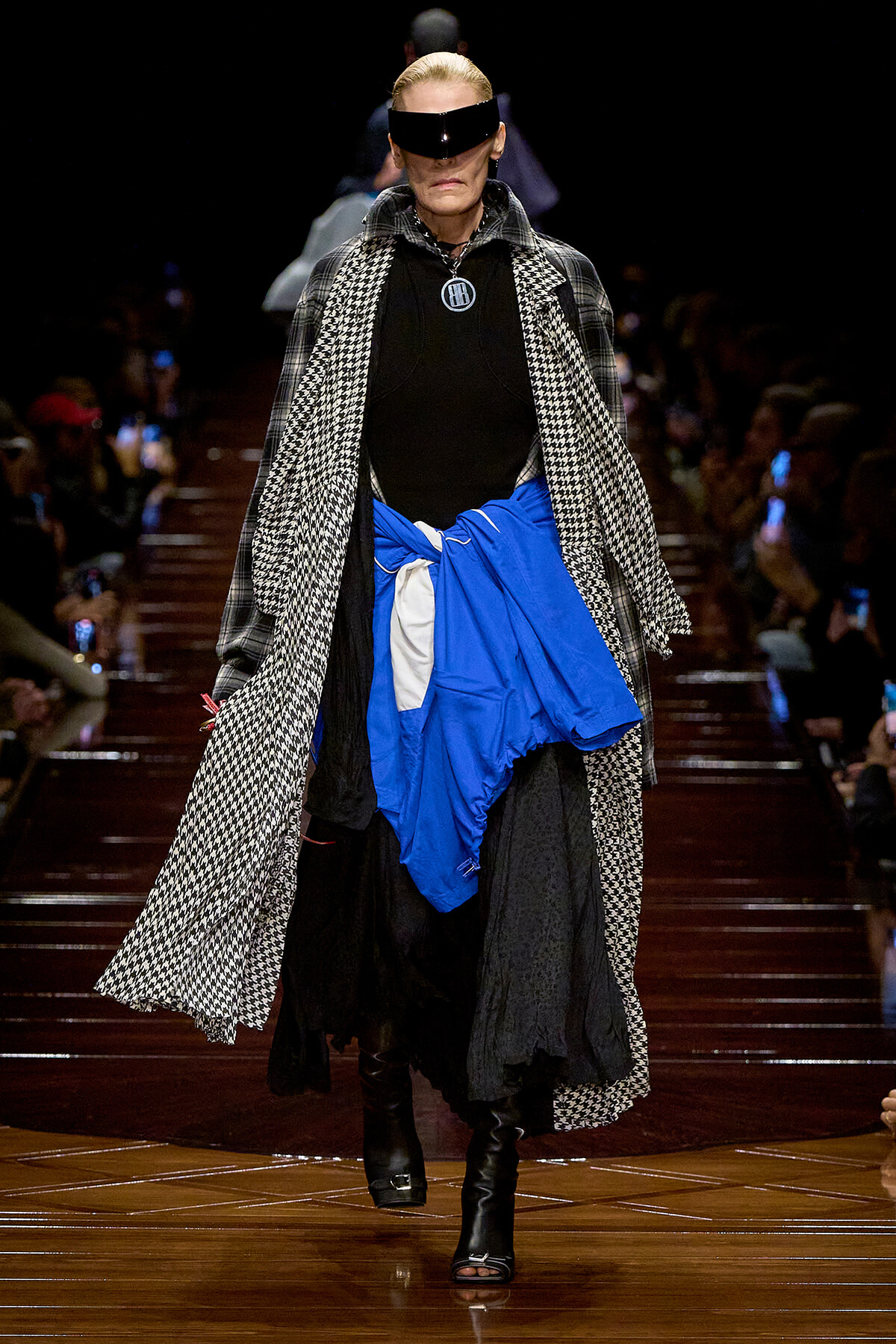 Model walks a fashion runway in a black outfit with a houndstooth coat, blue wrap skirt, and futuristic visor sunglasses.