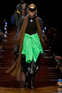 Model on a runway wearing a neon green jacket tied at the waist, a leopard-print scarf, plaid outerwear, black layers, and a futuristic visor.