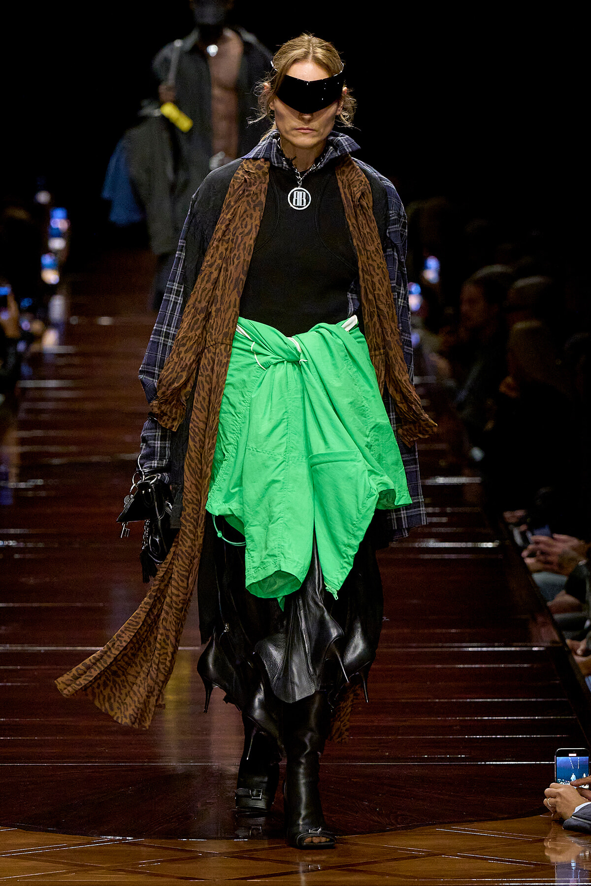 Model on a runway wearing a neon green jacket tied at the waist, a leopard-print scarf, plaid outerwear, black layers, and a futuristic visor.