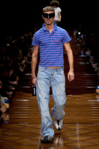 Male model walks a fashion runway in a blue striped polo, baggy light-wash jeans, metallic sneakers, and a wide black visor over his eyes.