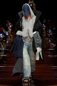 Model walking a runway in a gray zip hoodie under a black oversized coat, light-wash jeans, blue cap, and open-toe sandals on a glossy catwalk.