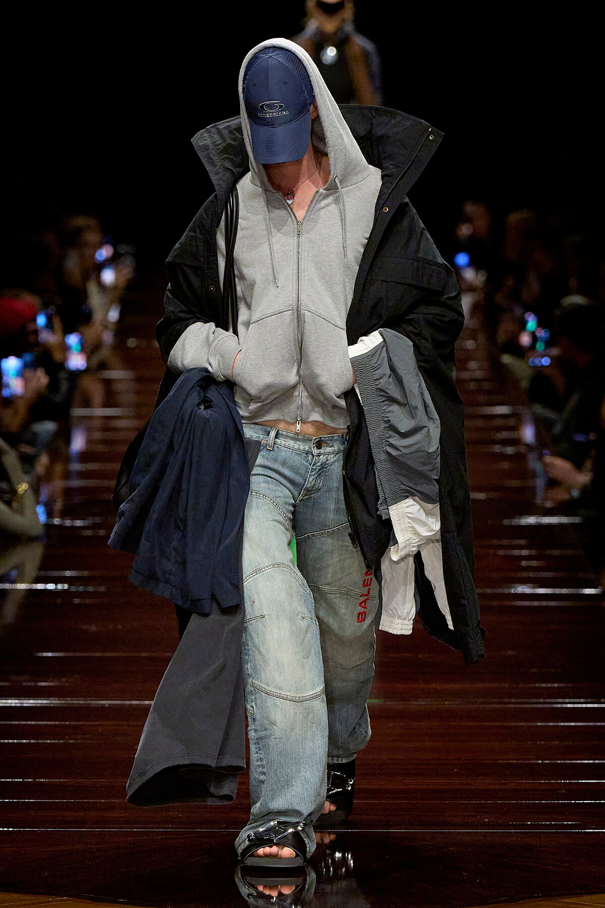 Model walking a runway in a gray zip hoodie under a black oversized coat, light-wash jeans, blue cap, and open-toe sandals on a glossy catwalk.