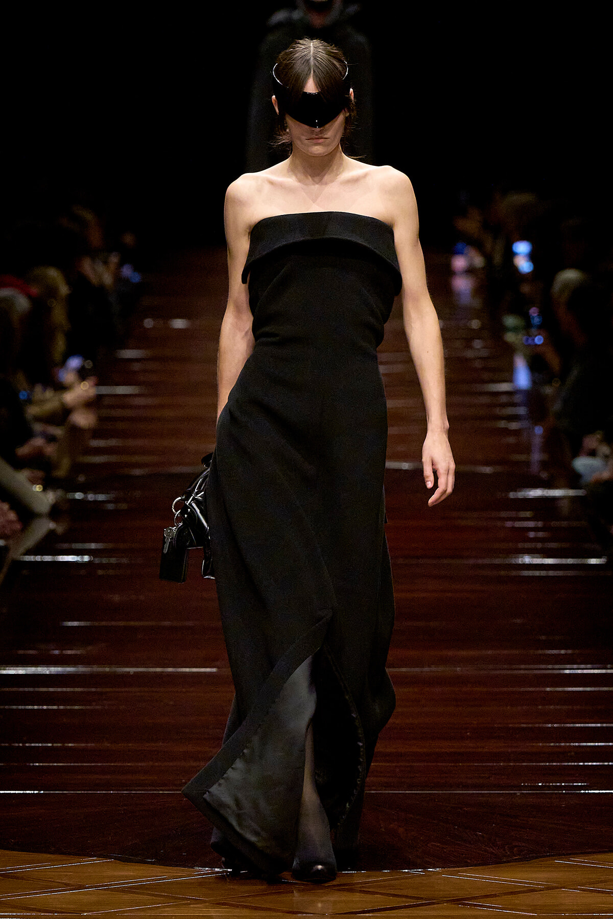 Model walks a runway in a black strapless gown, wearing a large black visor and carrying a small black handbag.