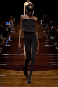 Model walking a fashion runway in an all-black strapless outfit with a blindfold and bold dangling earrings