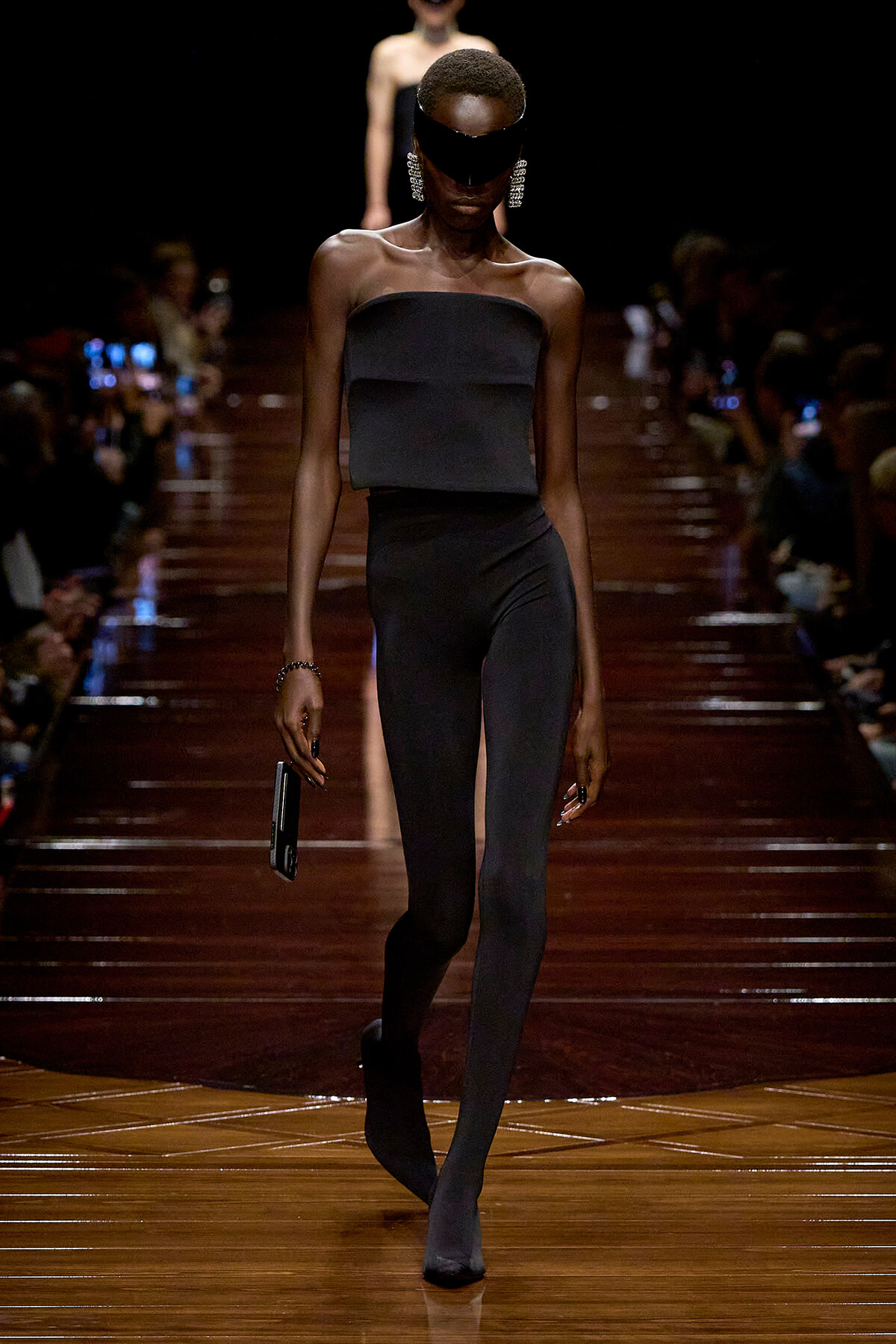Model walking a fashion runway in an all-black strapless outfit with a blindfold and bold dangling earrings