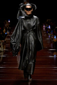 Model walking the runway in an oversized black leather coat with a large hood and wraparound visor sunglasses, holding a matching bag.