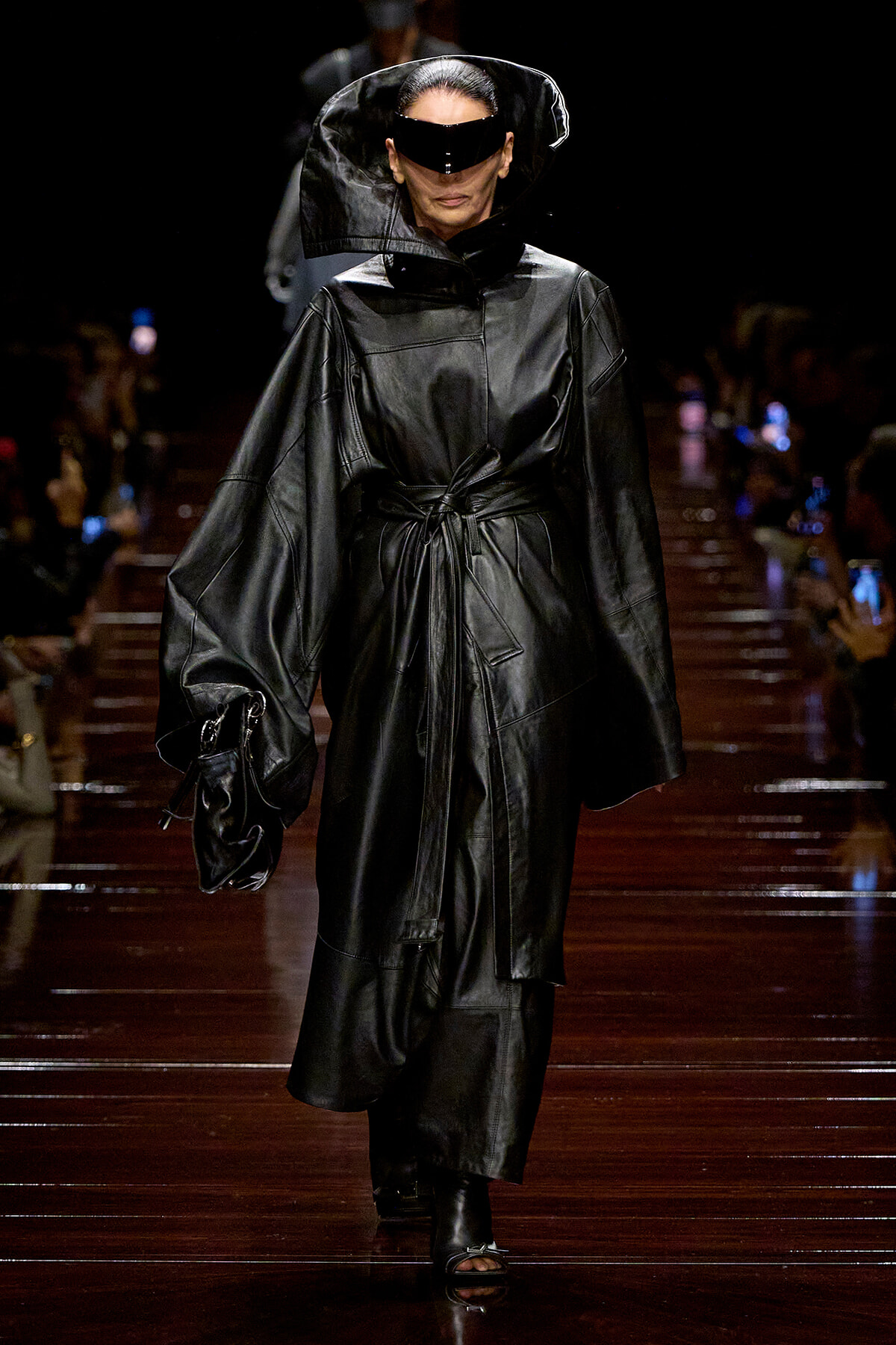 Model walking the runway in an oversized black leather coat with a large hood and wraparound visor sunglasses, holding a matching bag.