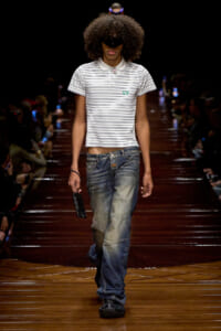 Male fashion model walking the runway in a white striped polo and oversized jeans, holding a black phone in his right hand with a large afro and sunglasses.