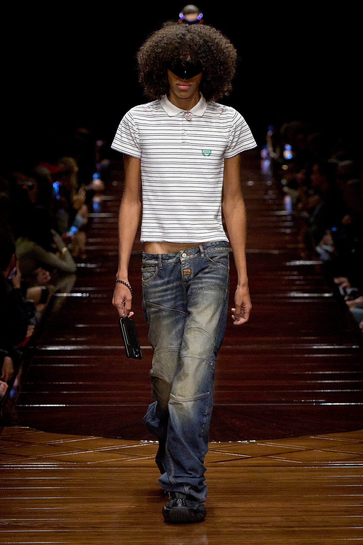 Male fashion model walking the runway in a white striped polo and oversized jeans, holding a black phone in his right hand with a large afro and sunglasses.