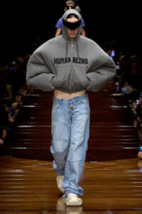 Model on a runway wearing a oversized gray hooded sweatshirt with 'HUMAN BEING' across the chest, baggy jeans, and white sneakers, hands in pockets.