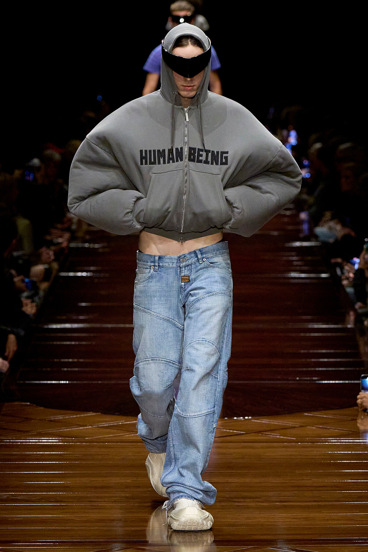 Model on a runway wearing a oversized gray hooded sweatshirt with 'HUMAN BEING' across the chest, baggy jeans, and white sneakers, hands in pockets.