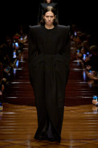 Model walking a runway in a long black double-breasted coat with oversized shoulders.