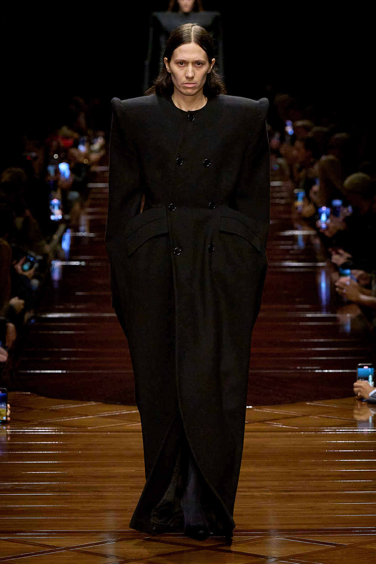 Model walking a runway in a long black double-breasted coat with oversized shoulders.