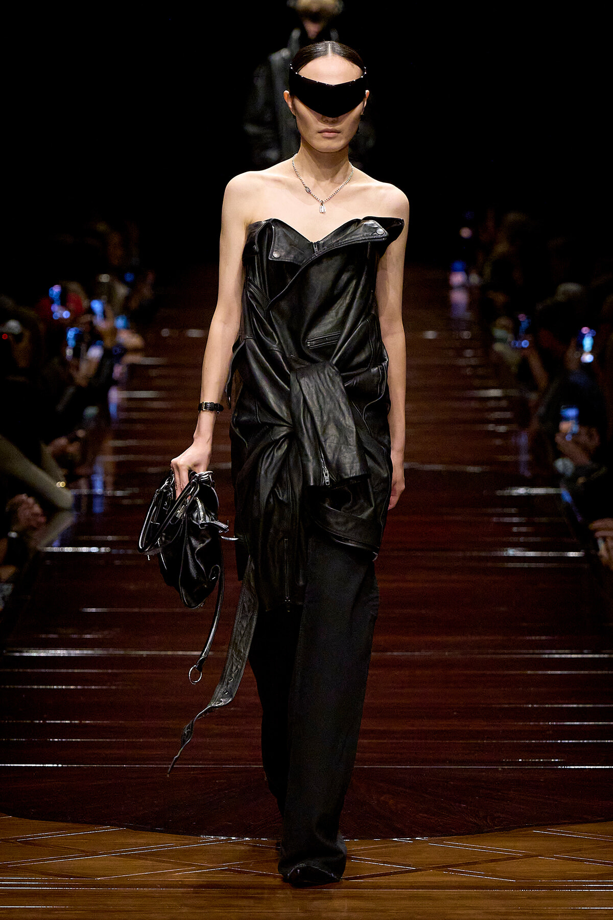 Model on a runway in a black leather off-shoulder outfit with a wide black visor, carrying a matching black handbag.