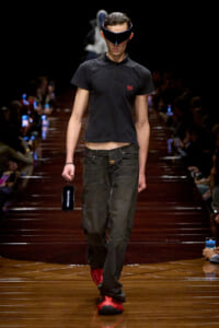 Male model on a fashion runway wearing a black polo, dark jeans, red sneakers, and a futuristic visor, holding a Balenciaga-branded phone.
