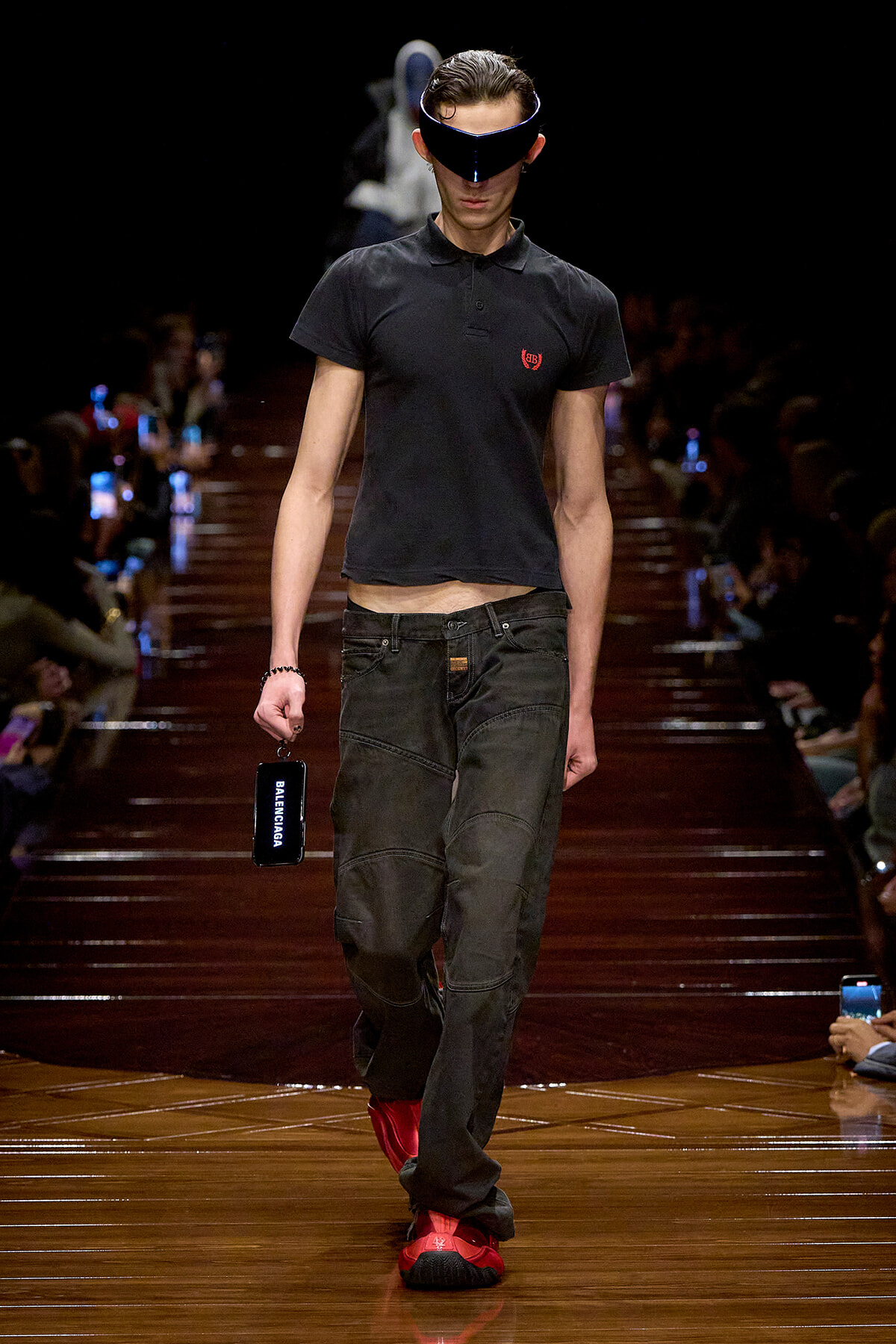 Male model on a fashion runway wearing a black polo, dark jeans, red sneakers, and a futuristic visor, holding a Balenciaga-branded phone.