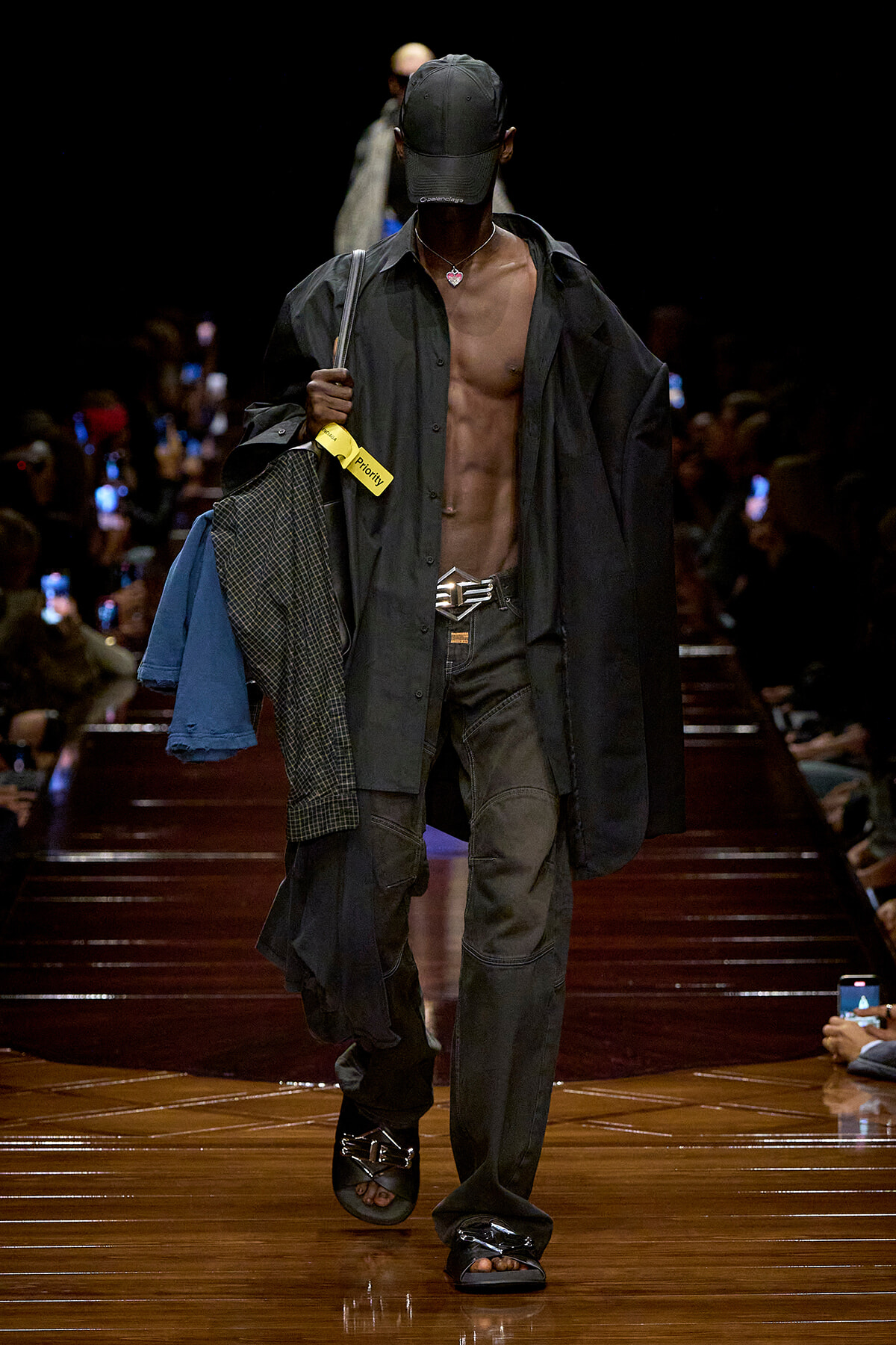 Male model walking a runway in an open black shirt, cap, and necklace, carrying clothes over one shoulder.