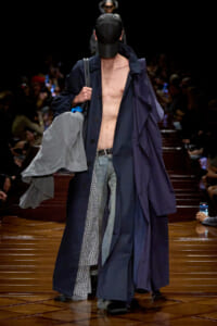 Male model on a runway wearing long navy trenches, cap, no shirt, jeans, and carrying a gray jacket over the shoulder.
