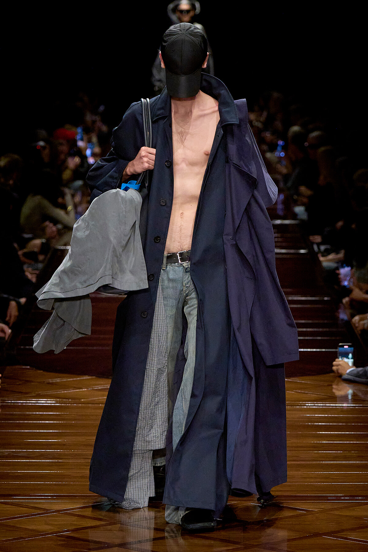 Male model on a runway wearing long navy trenches, cap, no shirt, jeans, and carrying a gray jacket over the shoulder.