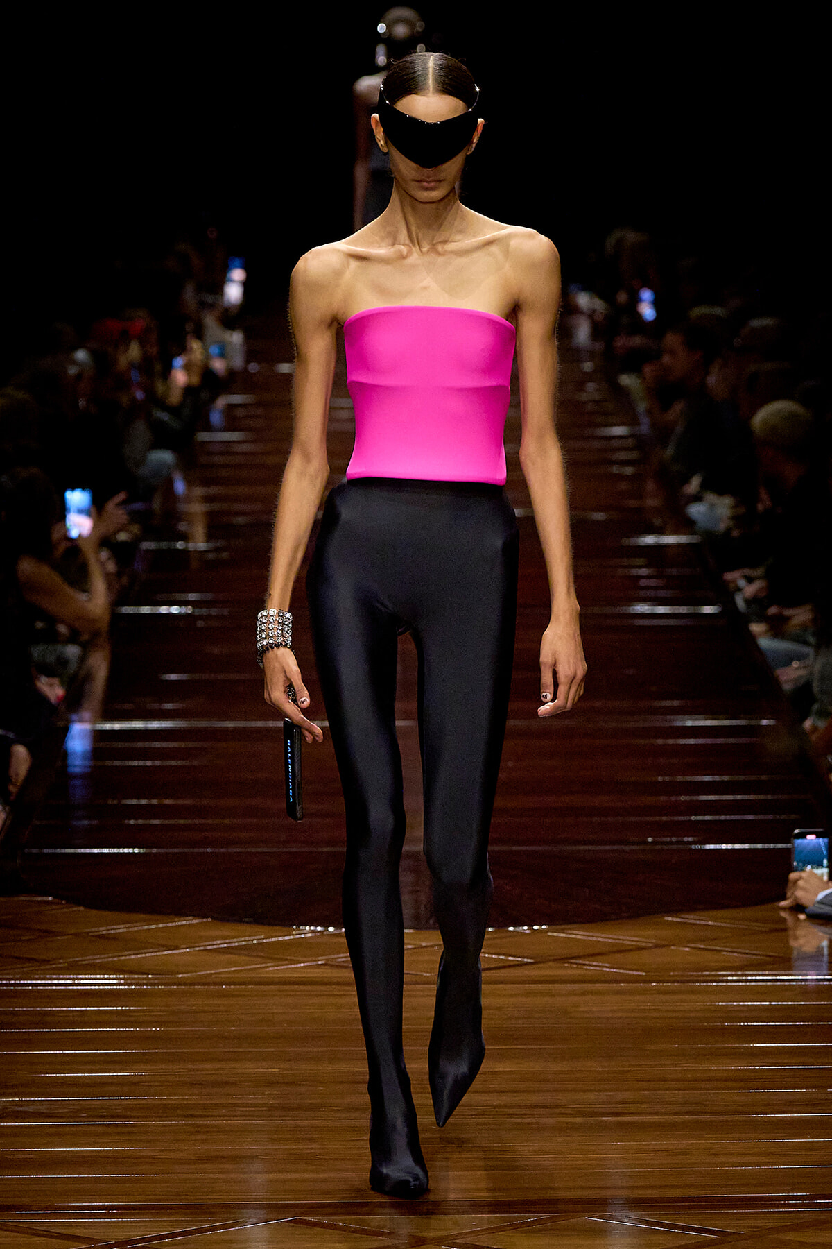 Model on a fashion runway wearing a bright pink strapless top and black glossy pants, with a black eye mask and chunky silver bracelet, holding a microphone.