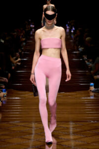 Model on a runway wearing a pink strapless crop top and matching high-waisted leggings, black visor sunglasses, and a metallic choker, holding a phone.