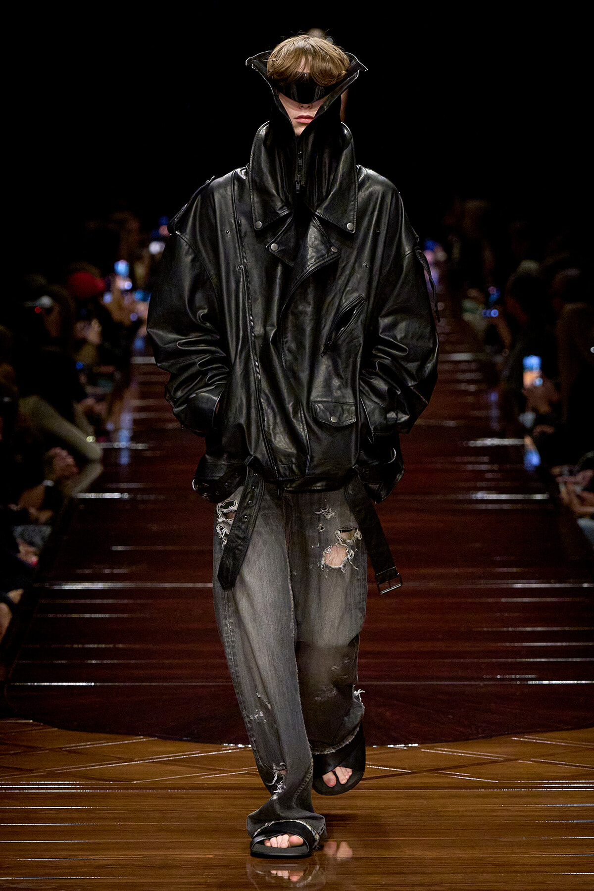 Model walks a fashion runway in an oversized black leather jacket with a high collar, distressed gray jeans, and open-toe sandals, wearing futuristic visor goggles.