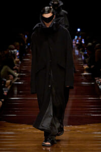 Model walks a dark runway in an oversized black coat with a hooded cape and futuristic black visor sunglasses, audience blurred in the background.