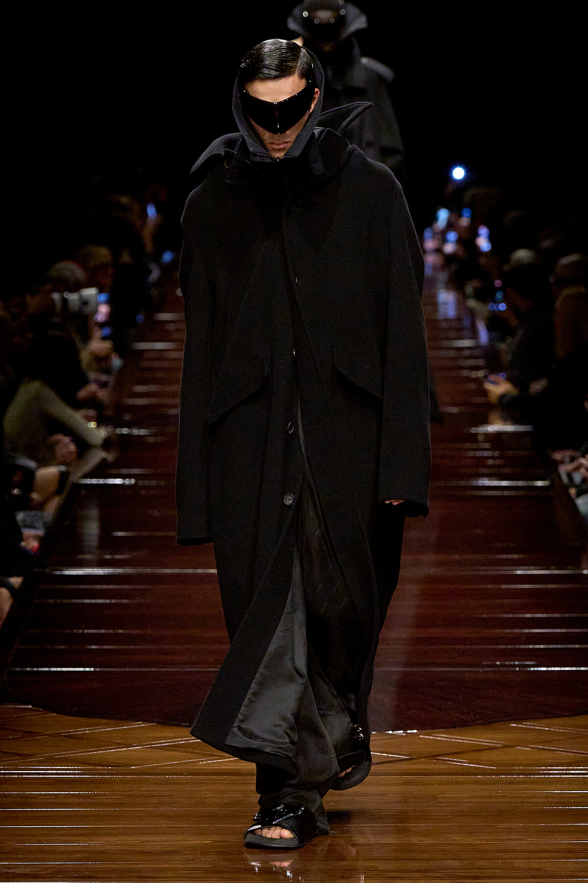 Model walks a dark runway in an oversized black coat with a hooded cape and futuristic black visor sunglasses, audience blurred in the background.