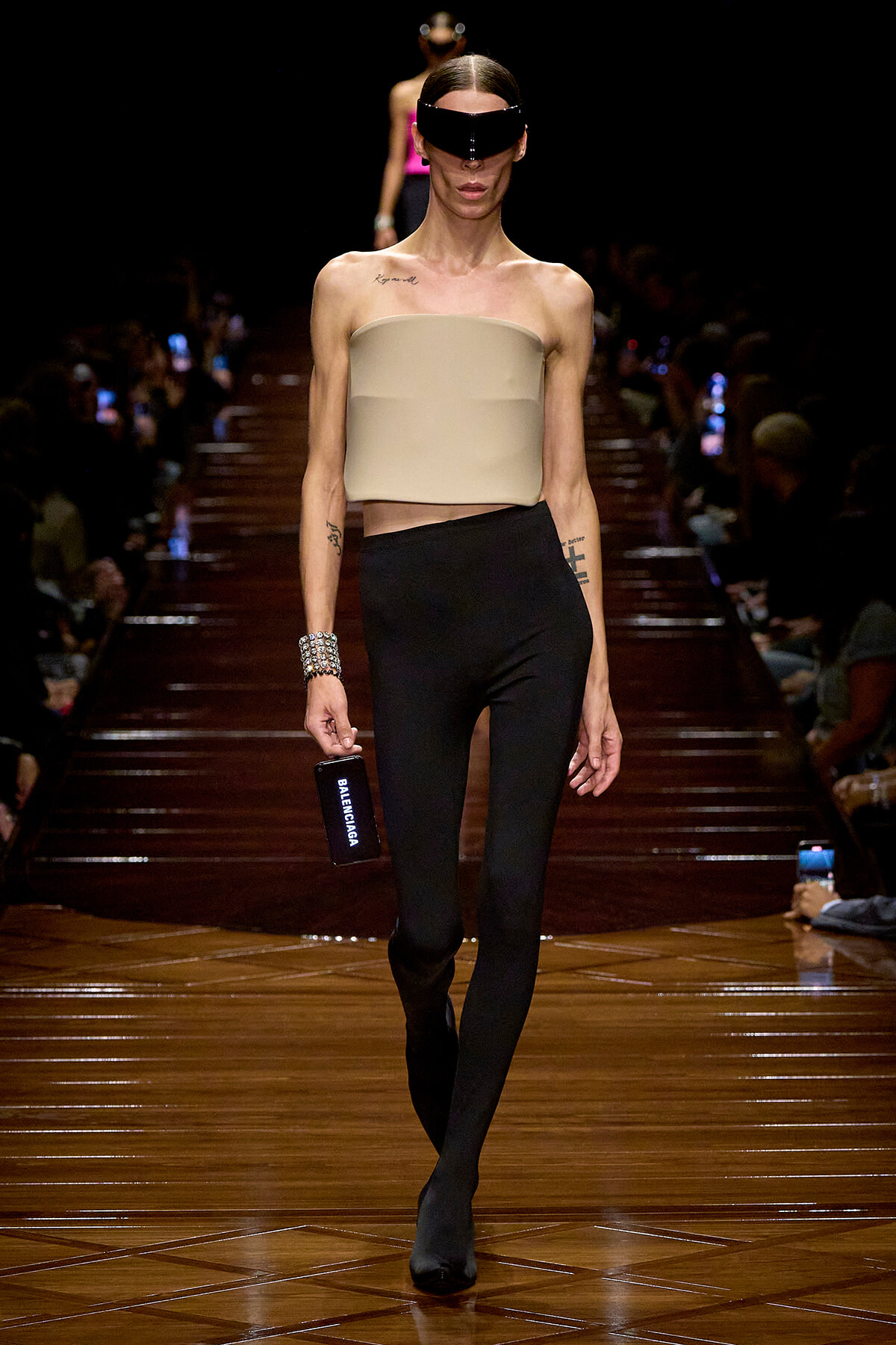 Model walks the runway in a beige bandeau top, black high-waisted leggings, and a futuristic black visor eyewear.
