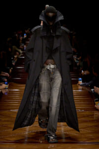 Model walking the runway in a long black trench coat over a dark outfit, oversized sunglasses, and unpacked jeans with a metallic belt; spotlighted on a glossy wooden catwalk.