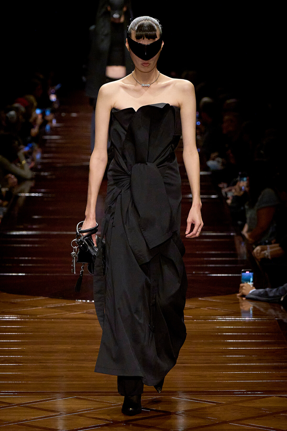 Model walks the runway in a strapless black gown with sculpted folds, paired with a black visor-style mask and a small handbag.