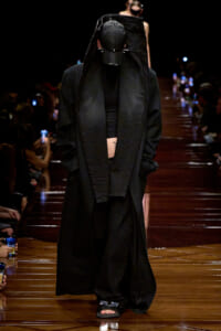 Model walks a black-on-black runway look: oversized coat, cap, and bare sandals, hands in pockets amid a dim audience.