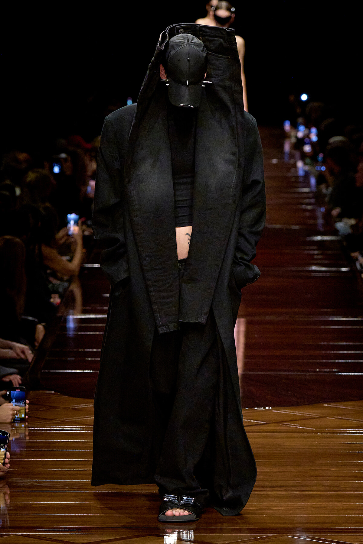 Model walks a black-on-black runway look: oversized coat, cap, and bare sandals, hands in pockets amid a dim audience.