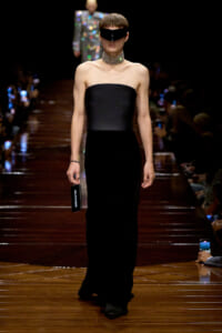 Runway model in a strapless black gown with a futuristic black visor and chunky choker, holding a Balenciaga phone as she walks the dark wooden catwalk.
