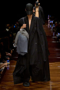 Model on runway wearing an oversized black coat and black cap, carrying a gray denim jacket over the arm and a shoulder bag; dark audience backdrop.