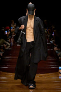 Male model walks a fashion runway in an all-black, oversized blazer and pants, cap, and sandals, carrying a black bag; torso is bare under the blazer.
