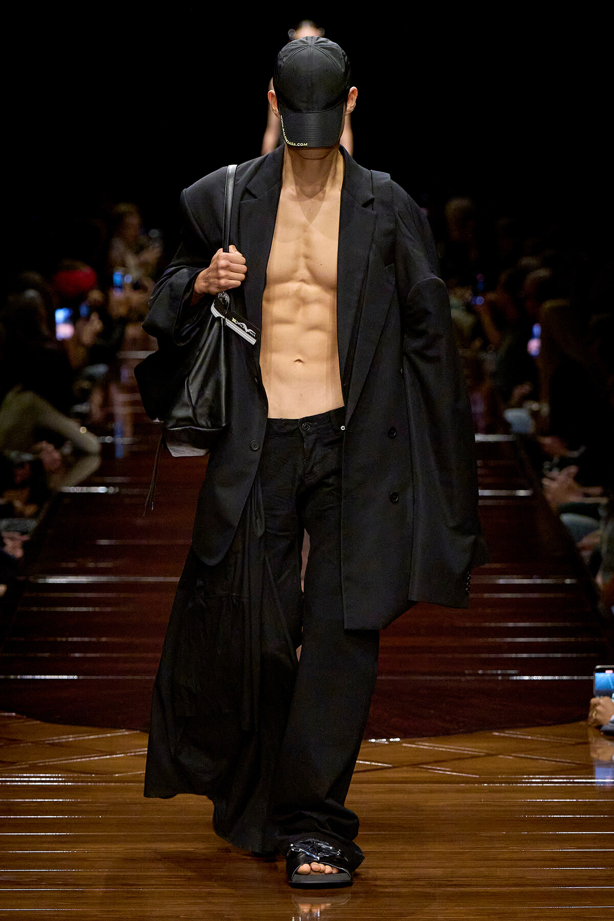 Male model walks a fashion runway in an all-black, oversized blazer and pants, cap, and sandals, carrying a black bag; torso is bare under the blazer.