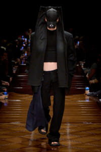 Fashion model walking a runway in an all-black outfit, oversized coat draped over head with cap, cropped top and loose black pants.