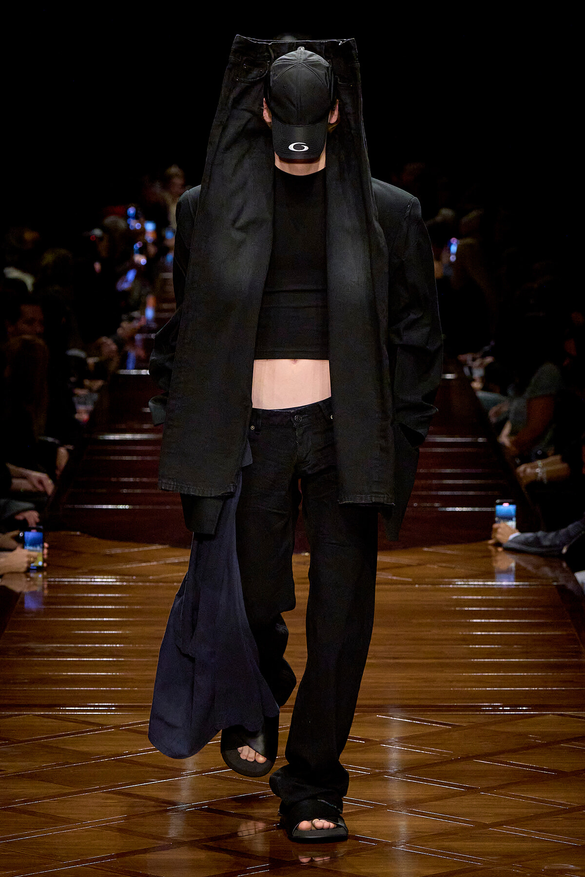 Fashion model walking a runway in an all-black outfit, oversized coat draped over head with cap, cropped top and loose black pants.