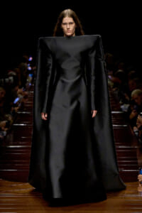 Model on a runway wearing a dramatic, oversized black gown with square shoulders and long sleeves.